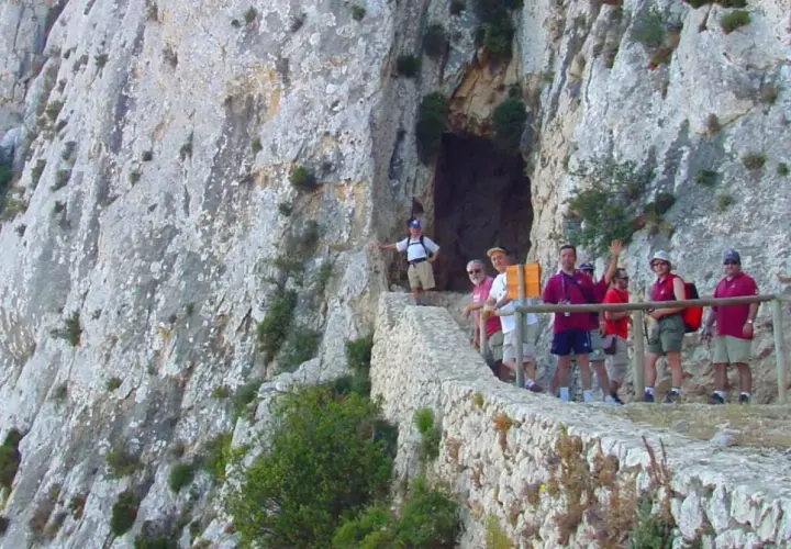 Grupo de excursionistas en el sendero del Peñón de Calpe, con una cueva a la izquierda y vistas rocosas.