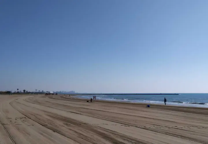 Amplia y tranquila playa de Meliana con arena marcada por vehículos. El mar azul se extiende hasta el horizonte, con algunas personas paseando.
