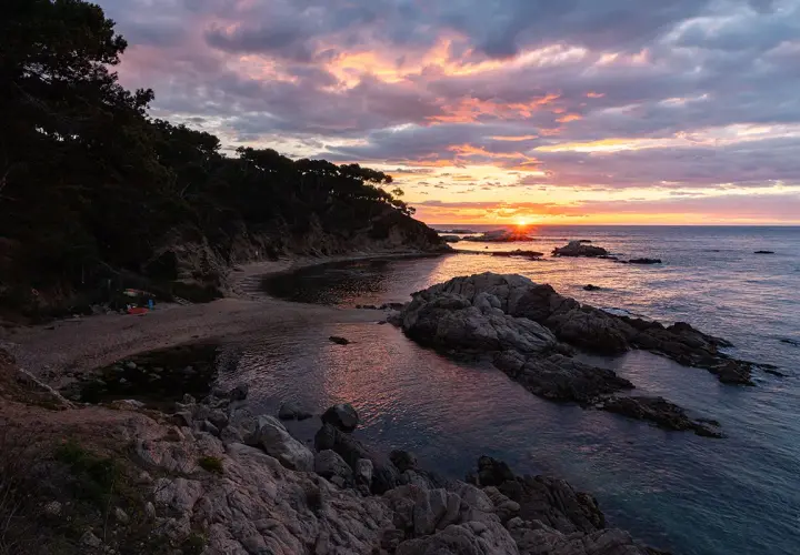 Atardecer en la punta estrella Moraira, con el sol asomando entre rocas y el mar reflejando los tonos rosados del cielo.