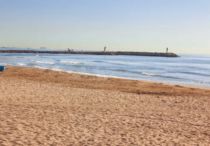 Arena dorada en la playa Port Saplaya, con el mar azul y un espigón con faros.