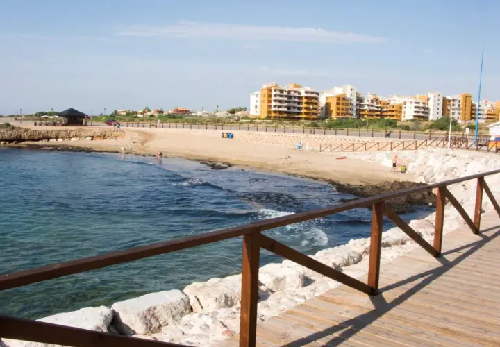Paseo de madera junto al mar en la cala Piteras. Edificios modernos y playa de arena dorada.