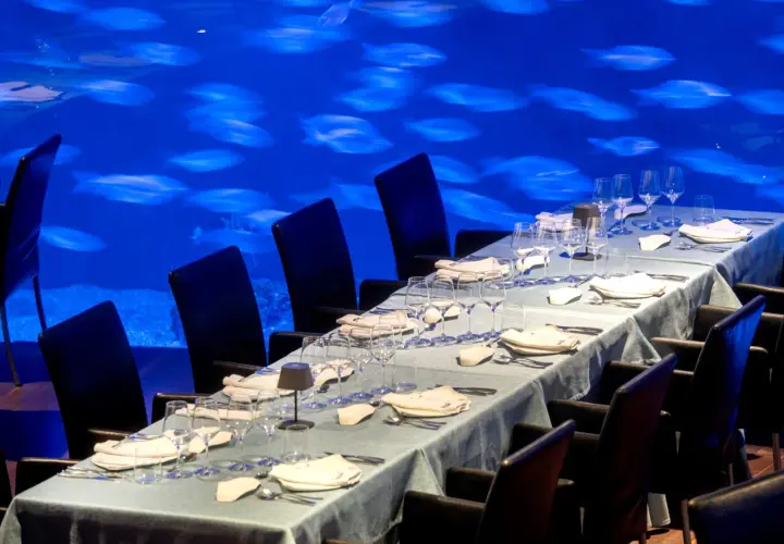 Mesa puesta para cena en el Oceanogràfic, con sillas negras y un acuario de peces de fondo. ¿Dónde comer en el Oceanogràfic? Opiniones.