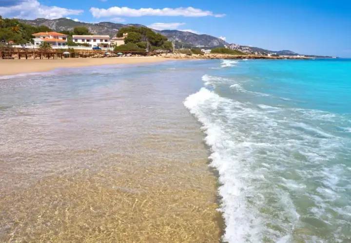 Aguas cristalinas y arena dorada en la playa la Romana, Alcossebre. Un paraíso mediterráneo con montañas al fondo.