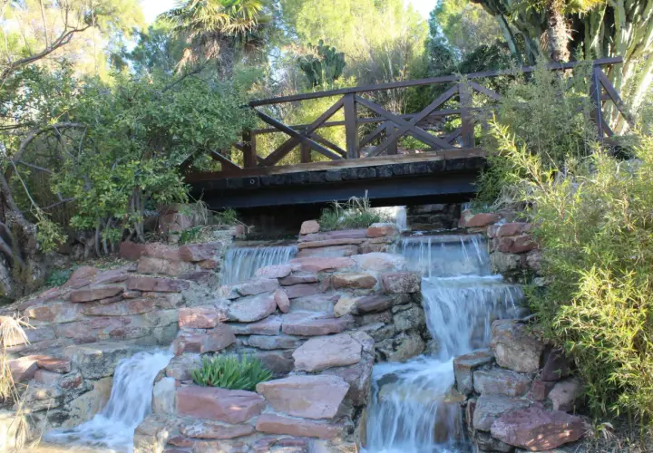Puente de madera sobre cascadas en el parque de Benicalap, rodeado de vegetación exuberante y rocas rojizas.