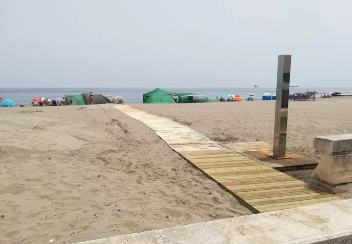 Pasarela de madera hacia las playas con duchas en Alicante. Gente disfrutando del sol bajo sombrillas y tiendas verdes.