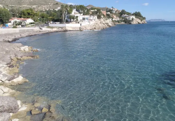Aguas cristalinas bañan la costa rocosa de cala Piteres, con casas blancas y palmeras en la ladera.