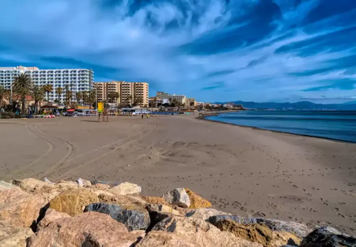 Amplia playa de arena con edificios hoteleros al fondo. El mar azul y el cielo nublado crean un paisaje sereno, ideal para pasear y sentir la **playa fuente de la salud**.