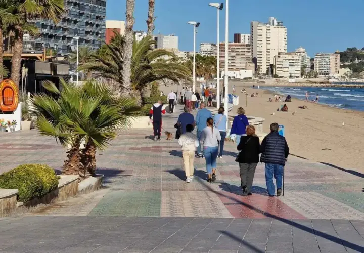 Gente paseando por el **paseo marítimo de Calpe**, con palmeras, edificios altos y la playa al fondo.