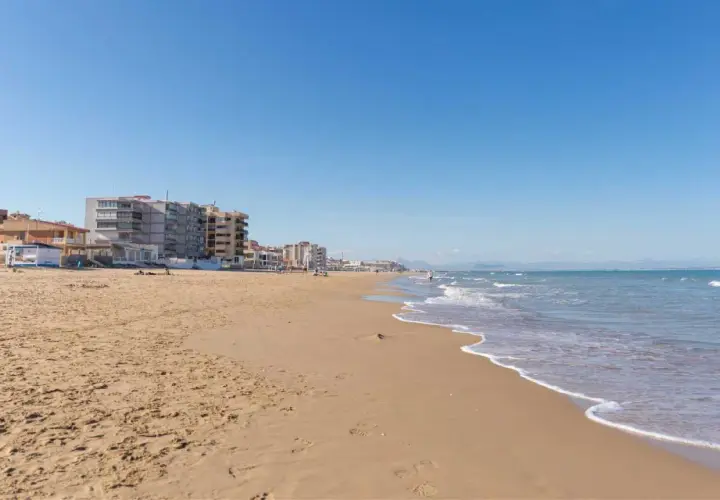 Arena dorada y mar azul en la playa La Roqueta, con edificios de apartamentos y montañas al fondo.