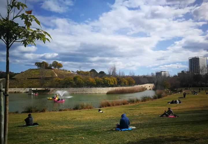 Gente relajándose en el césped del parque de cabecera, con un lago y barcas de pedales al fondo.