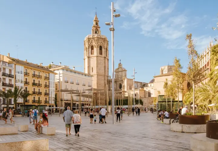 Gente pasea por la plaza frente a la Catedral de Valencia, con el icónico campanario del Miguelete dominando el cielo azul.