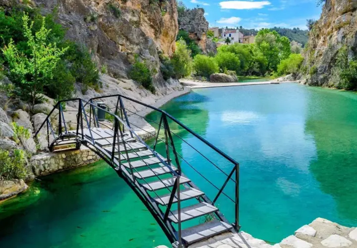 Puente metálico sobre las aguas turquesas del **río Bolbaite**, rodeado de rocas y vegetación exuberante.