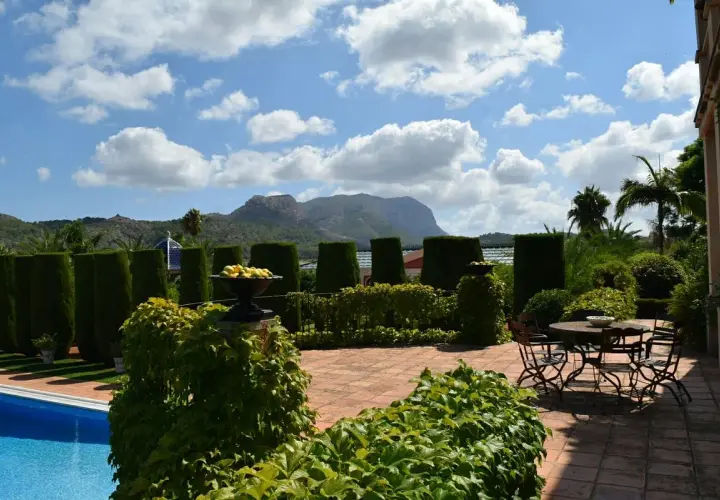 El jardín de l'albarda, con piscina azul, setos verdes, mesa y sillas, y montañas al fondo bajo un cielo nublado.