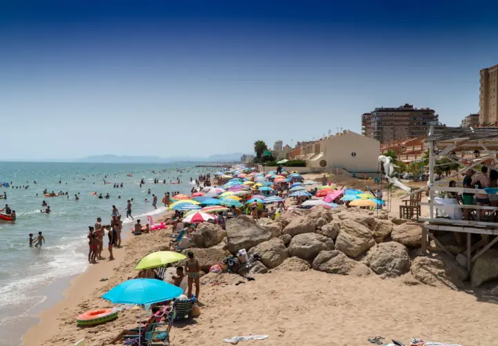 Día soleado en la playa Tavernes de la Valldigna, con mucha gente disfrutando del mar y la arena bajo coloridas sombrillas.
