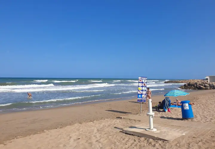 Un día soleado en la playa el Marenyet. Olas rompen en la orilla mientras una persona se baña. Hay un cartel de normas y una sombrilla.