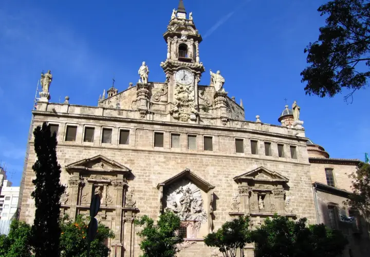La **iglesia de los Santos Juanes**, con su imponente fachada de piedra, torre con reloj y estatuas, se alza majestuosa bajo un cielo azul intenso.
