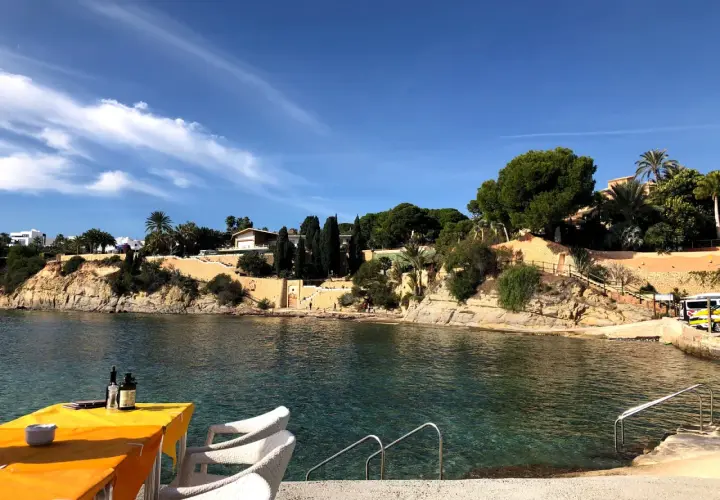 Mesa con mantel naranja y sillas blancas junto al mar cristalino en cala les Bassetes. Casas y vegetación en la ladera.