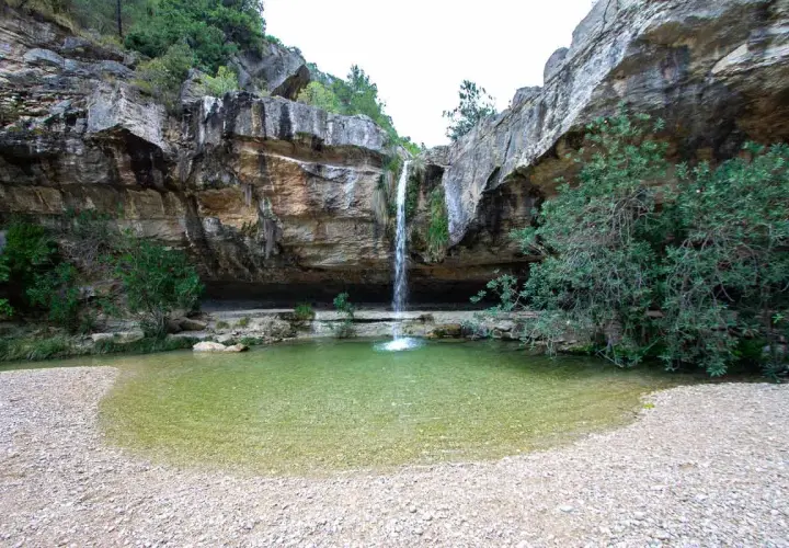 Cascada cayendo en **charcos de quesa** de agua cristalina, rodeada de rocas y vegetación exuberante.