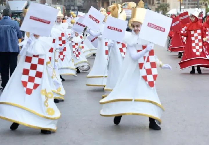 Participantes en la cabalgata del ninot, con trajes blancos y rojos, portan carteles con palabras como "Ciencia" y "Educació".