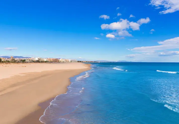 Amplia playa de arena dorada en Sagunto, con el mar Mediterráneo azul y edificios de la costa al fondo bajo un cielo despejado.