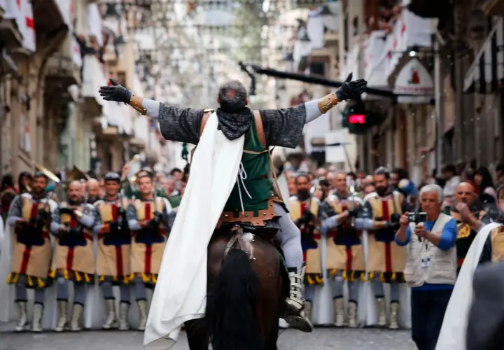 Un jinete con capa blanca lidera un desfile de moros y cristianos, con gente observando y banderas en las calles.