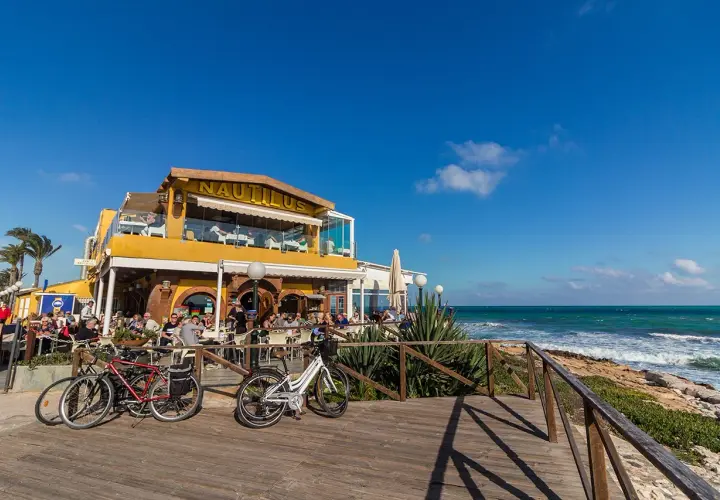 Restaurante Nautilus en la playa Punta Prima, con gente disfrutando en terrazas y bicicletas aparcadas.