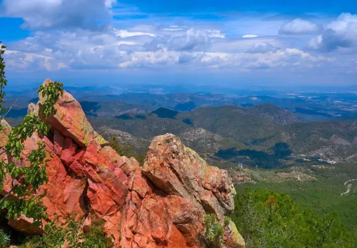 Rocas rojizas y vegetación verde en primer plano, con un vasto paisaje montañoso de la sierra de Espadán bajo un cielo azul con nubes.