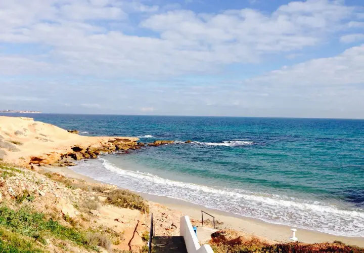 Escaleras descienden a la playa rocamar, con arena dorada, rocas y mar azul bajo un cielo nublado.