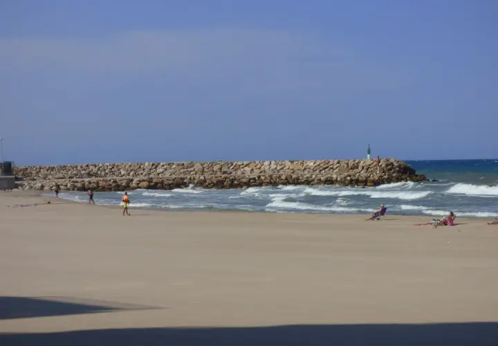 Una playa tranquila con una rompeolas de rocas. Gente relajándose en sillas de playa y paseando. El mar azul y el cielo despejado crean una escena de verano perfecta.