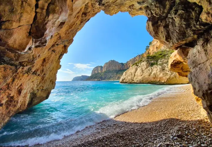 Vista desde una cueva hacia la cala Testos en Benitachell. Aguas turquesas, arena de guijarros y acantilados dorados bajo un cielo azul.