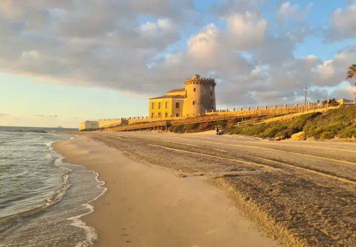 Atardecer en la playa del Conde, con una torre antigua y un paseo de madera. El mar acaricia la arena dorada bajo un cielo nublado.