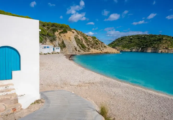 Casa blanca con puerta azul en la playa de guijarros de Vall d'Ebo, con aguas turquesas y colinas verdes.