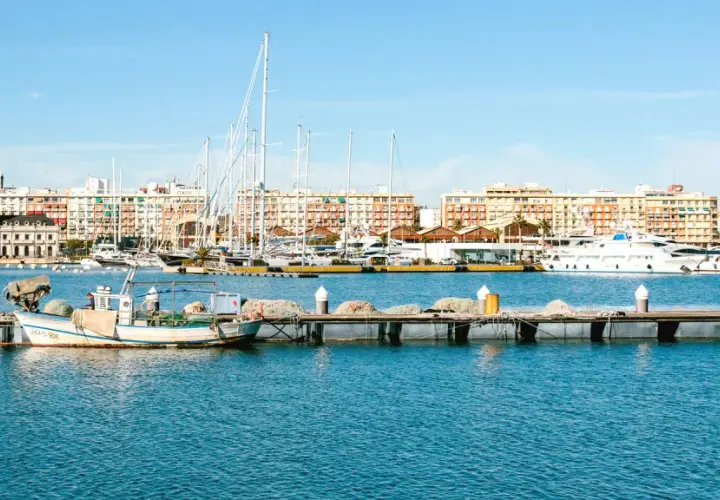 Barcos de pesca y yates de lujo atracados en la marina de Valencia, con edificios coloridos de fondo bajo un cielo azul.
