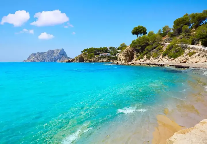 Aguas turquesas y arena dorada en la cala Pinets, con rocas y pinos coronando la costa. Un paraíso mediterráneo.
