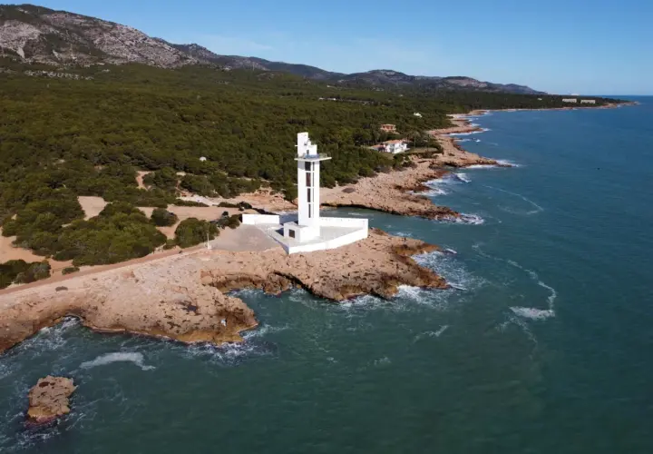 Faro blanco moderno en la costa rocosa de la Sierra de Irta, con el mar azul y montañas verdes al fondo.