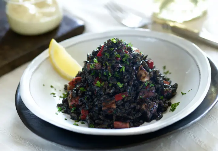 Delicioso arroz negro con trozos de calamar y pimiento rojo, adornado con perejil fresco y una rodaja de limón.