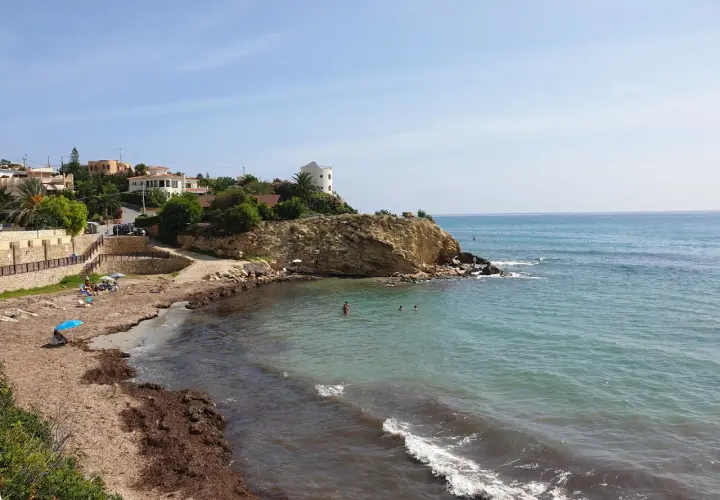 La cala Coveta, con su playa de arena y rocas, invita a disfrutar del mar Mediterráneo. Casas blancas coronan la colina.
