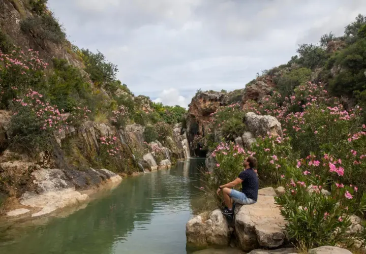 Hombre sentado junto a un río en un cañón con flores rosas. Un lugar perfecto para excursiones cerca de Valencia.
