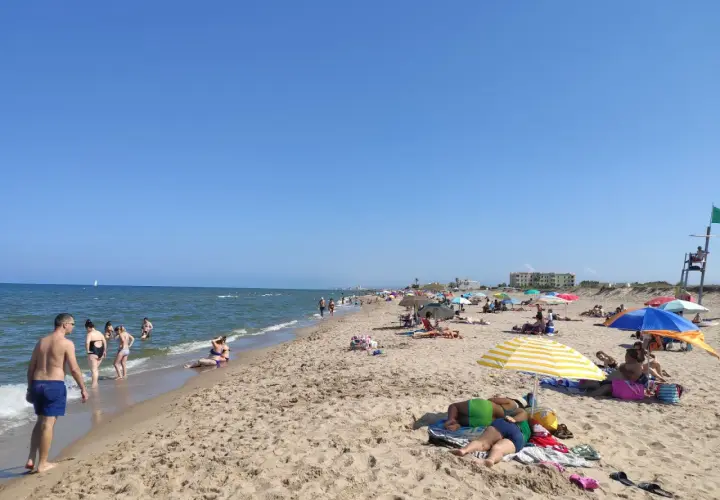 Gente disfrutando del sol y el mar en la playa de la Garrofera. Sombrillas, toallas y bañistas llenan la arena dorada bajo un cielo azul.