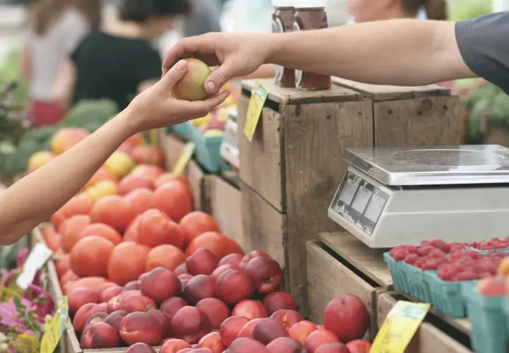 Manos intercambian una manzana en un mercadillo de oliva. Tomates, frambuesas y melocotones esperan ser pesados.