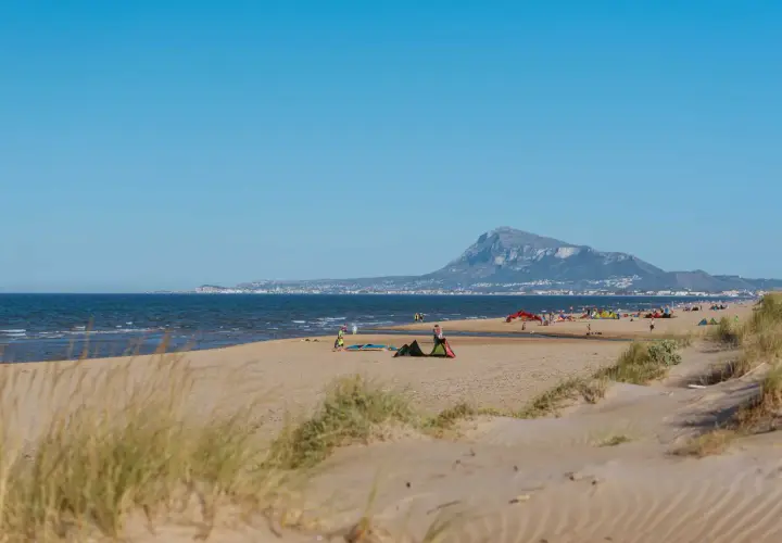 Dunas y mar en la playa de Rabdells, con gente disfrutando del sol y el paisaje montañoso al fondo.