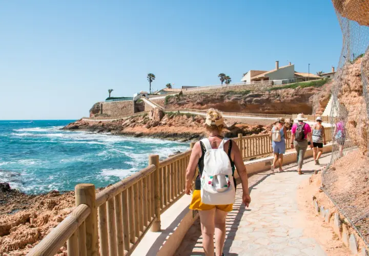 Paseo por la costa en cala capitan. Turistas caminan por un sendero con vistas al mar azul y acantilados rojizos.