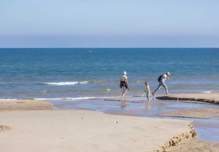 Un niño y dos adultos disfrutan de un día soleado en la playa Pau Pi, jugando cerca de la orilla del mar azul.