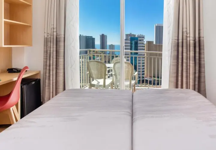 Habitación de hotel pueblo Benidorm con vistas al mar y rascacielos. Balcón con mesa y sillas. Escritorio con silla roja y minibar.