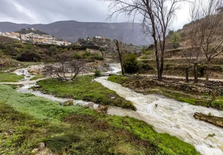 Río caudaloso cerca de Sella, Alicante, con cascadas y un pueblo blanco en la ladera.