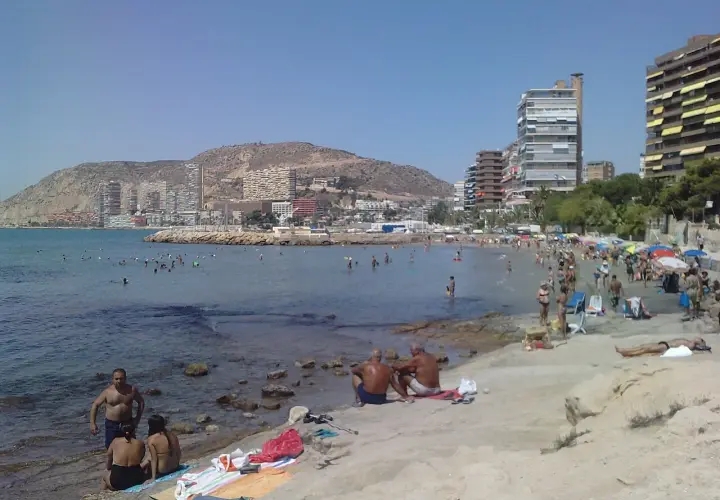 Gente disfrutando del sol y el mar en la playa de la Almadraba, con edificios modernos al fondo.