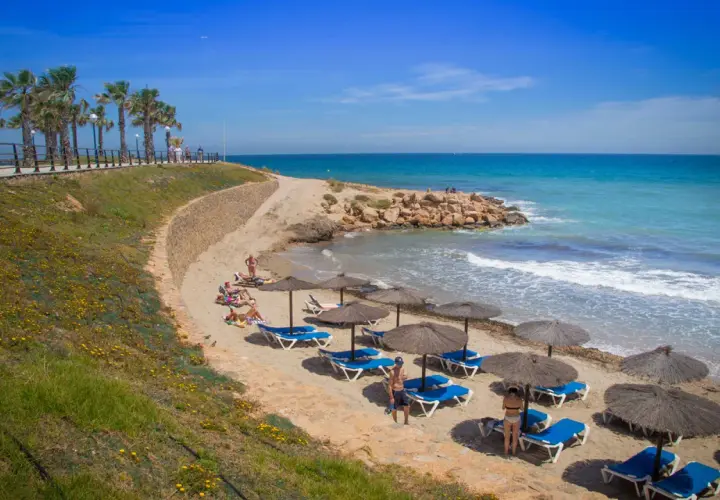 Una cala estaca con palmeras y sombrillas en la arena, donde la gente descansa bajo el sol. El mar azul turquesa invita a un refrescante baño.