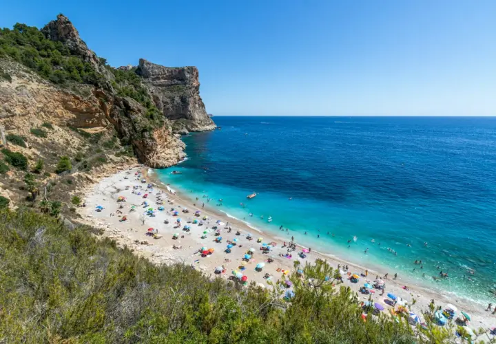 La **cala Moraig** es un paraíso de guijarros y aguas turquesas, con acantilados imponentes y gente disfrutando del sol bajo sombrillas de colores.