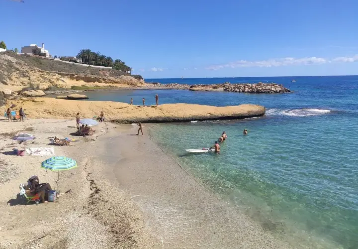 Gente disfrutando del sol y el mar en la cala Almadraba, Campello. Aguas cristalinas y rocas emergiendo del azul.