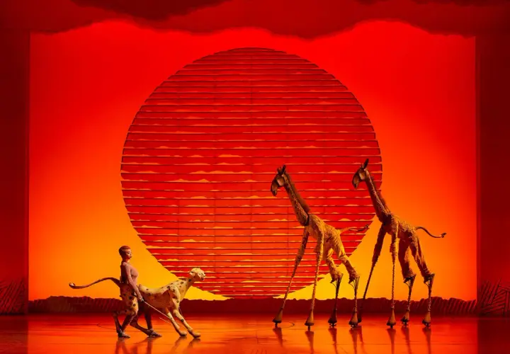Escena de teatro en Valencia con un guepardo y jirafas con zancos ante un sol naranja.